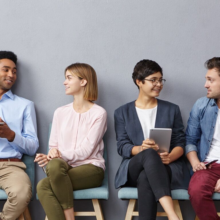 Horizontal portrait of people sit in queue, have pleasant conversation with each other, share ideas and life experience, isolated over grey concrete wall. Diverse group in row, speak and hold gadgets