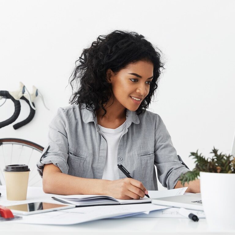 Happy attractive experienced African American female designer writing down some measured data in her notebook from laptop pc, with electronic devices and drawings on desk and bicycle in background